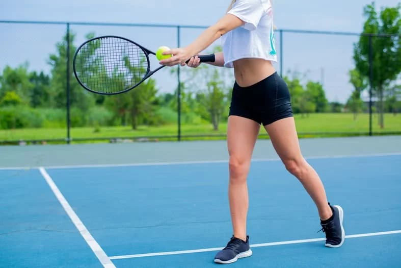 a picture of a woman's body from waist dwon holding a tennis racket with a green ball on a tennis court
